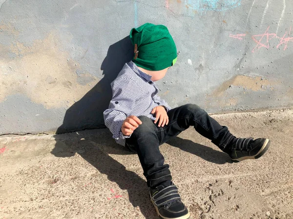 Een ongehoorzaam kind zit op straat bij de muur en draait zich om. Rechtenvrije Stockafbeeldingen
