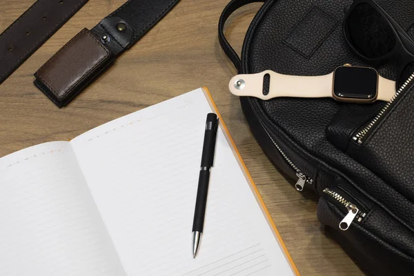 Open notebook with a black pen, smart watch and sunglasses on a bag and a leather belt on a wooden background. Home office concept