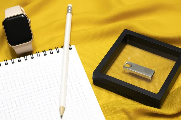 Damalı bir not defterinde beyaz bir kalem, akıllı bir saat ve yeni bir flaş belleğin yanında. Notlar için boş alan. Düz yatar, sarı arkaplan.