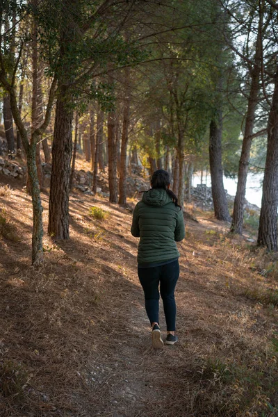 Caucasian woman with green jacket walking on a path between pine trees and a lake on a sunny day
