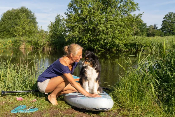Tişört ve şort giymiş bronzlaşmış bir kadın. Yelkenli için yiyecek panosunu hazırlıyor. Alman çoban köpeği kürek tahtasında oturuyor. Hayvan teması, spor ve eğlence. Rıhtım, kamış, nehir, mutlu.