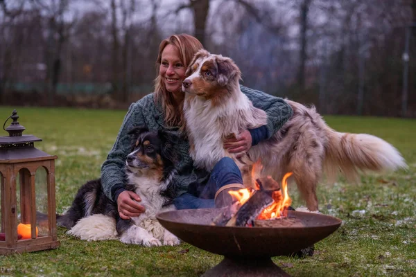 Young woman is sitting outside in the woods with her two Australian Shepherd dogs. Snow on the grass, twilight by the campfire Stock Picture