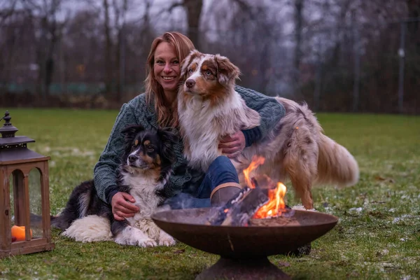 La joven está sentada afuera en el bosque con sus dos perros pastor australianos. Nieve en la hierba, crepúsculo junto a la fogata. Acurruces y animales de compañía — Foto de Stock