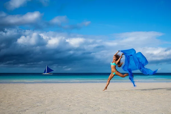 Beautiful young woman jumping on the beach with a blue tissue — Stock Photo, Image