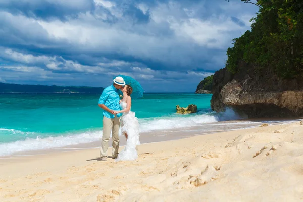 Bride and groom with blue umbrella kiss on the tropical coast — Stock Photo, Image