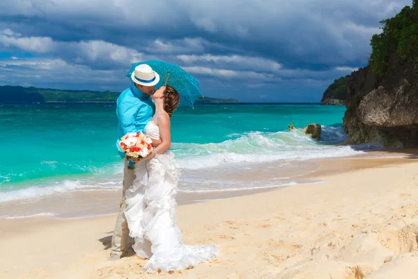 Bride and groom with blue umbrella kiss on the tropical coast — Stock Photo, Image