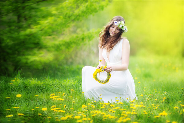 Image of pretty woman in a white dress weaves garland from dandelions in the field, happy cheerful girl resting on dandelions meadow, relaxation outdoor in springtime, vacation.