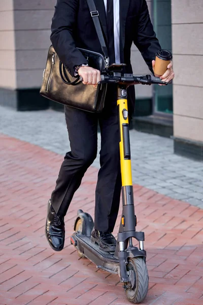 Cropped man in suit riding an electric scooter going at business meeting, holding coffee