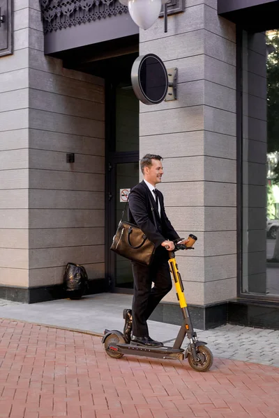 Modern elegant businessman in suit moving on electric scooter along exterior of office center — Stock Photo, Image