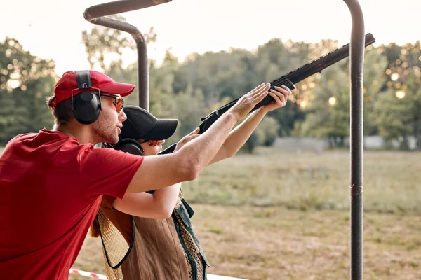 Homem caucasiano agradável no fone de ouvido está ensinando feminino a usar rifle, arma ao alcance — Fotografia de Stock