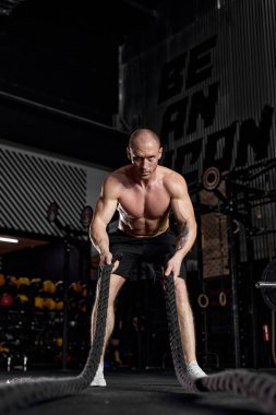 Üstsüz, beyaz bir adam. Çapraz formda spor salonunda savaş halatlarıyla çalışıyor..