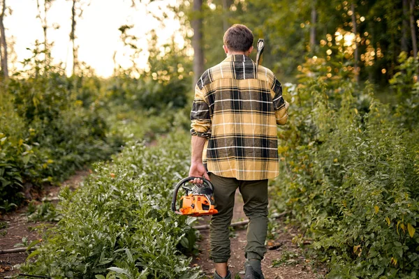 Vista desde atrás El joven leñador que va al bosque con motosierra para cortar árboles —  Fotos de Stock