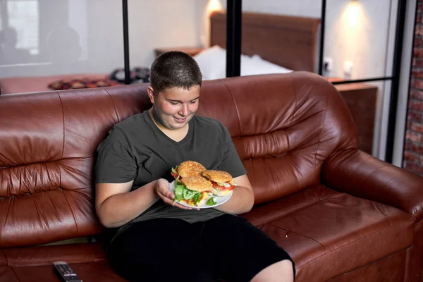 Feliz menino com sobrepeso sentar segurando prato com hambúrgueres no sofá de couro na sala de estar — Fotografia de Stock