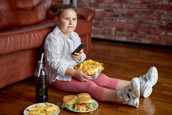 Vet meisje eten chips uit kom terwijl zitten op de vloer in de woonkamer, zijaanzicht — Stockfoto