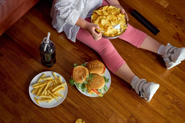 Tidak dikenal gadis makan keripik kentang renyah lezat dan makanan cepat saji duduk di lantai — Stok Foto