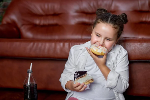 Avida ragazza si sente fame e mangiare ciambelle dolci mentre seduto sul pavimento a guardare la tv — Foto Stock