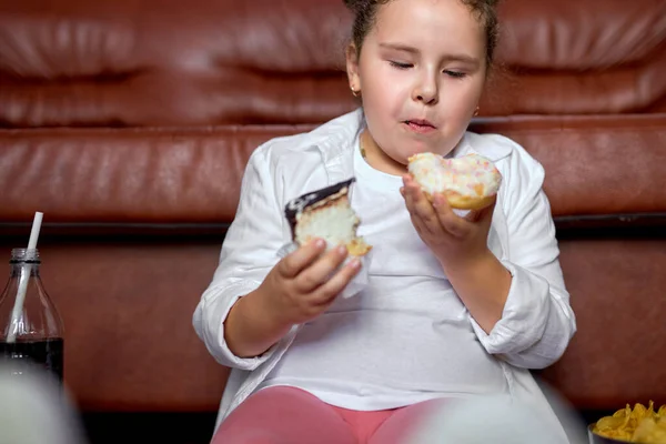 Caucasico ragazza sta mangiando malsano dolci torte e guardare la tv seduto sul pavimento. — Foto Stock