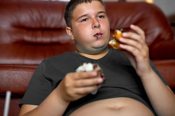 Il ragazzo affamato sta mangiando cibo spazzatura a casa mentre guarda la TV. Fast food malsano. — Foto Stock