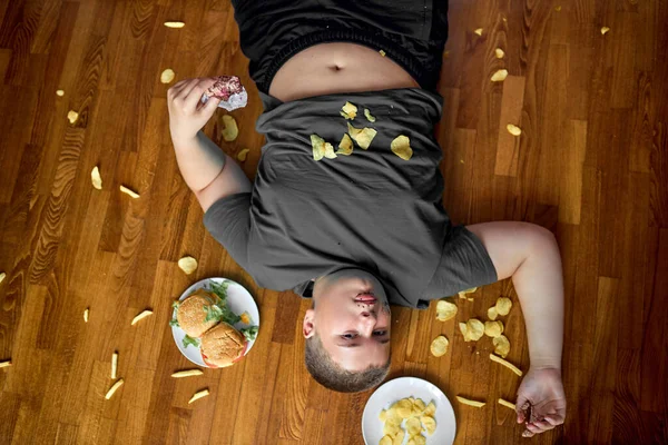 Sovrappeso grasso ragazzo mangiare cibo spazzatura mentre sdraiato sul pavimento a casa, con patatine fritte — Foto Stock
