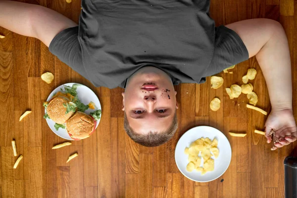 Vista dall'alto su grasso ragazzo in sovrappeso sdraiato sul pavimento circondato da cibo spazzatura, patatine fritte e patatine — Foto Stock