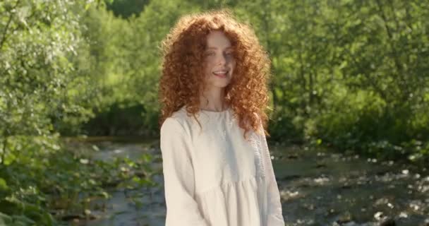 Wanita berambut merah yang menarik dengan gaun romantis putih menatap kamera, dekat danau — Stok Video