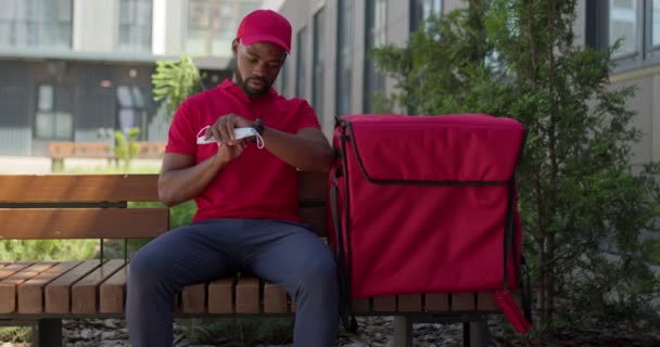Zwarte levering man controleren van de tijd, het dragen van masker en ga naar klanten — Stockvideo