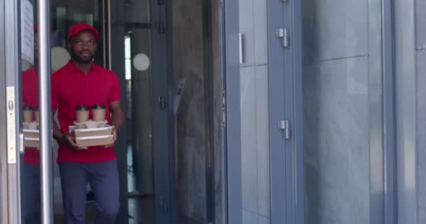 Barbudo africano repartidor en uniforme rojo con comida loncheras y café — Vídeos de Stock