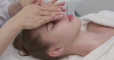 Kozmetik uzmanı yüz masajı yaparken rahat genç bayan kanepede dinleniyor.