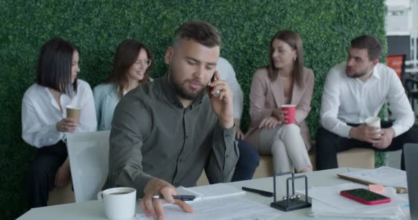 Кавказский человек разговаривает по смартфону в офисе, пока коллеги делают перерыв — стоковое видео