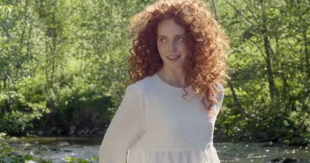 Belle femme aux cheveux rouges en robe romantique blanche regardant la caméra, près du lac — Video