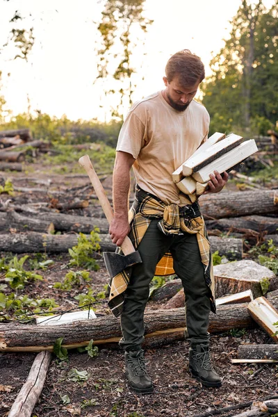 Fit unshaven guy in T-shirt holds firewood in hands at summer in forest. — Stock Photo, Image