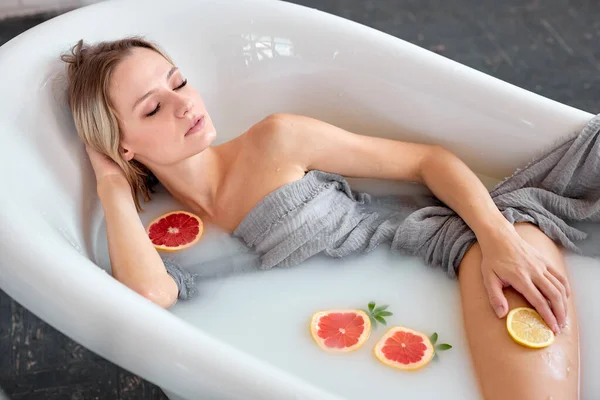 Приваблива жінка відпочиває у ванні, наповненій водою з молоком з цитрусами та квітами — стокове фото