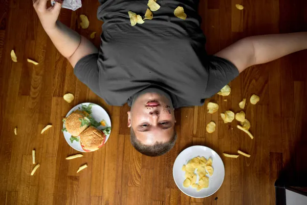 Vista dall'alto sul ragazzo mangiare troppo sdraiato sul pavimento in soggiorno, giovane ragazzo adolescente caucasico — Foto Stock