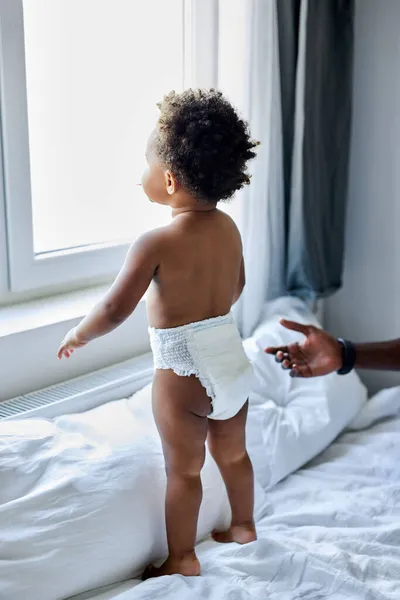 Curioso negro de piel oscura niña de pie en la cama mirando a través de la ventana, interesado — Foto de Stock