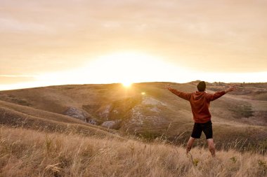 Adam, elleri havada, dağ manzarasının arka planında duruyor..
