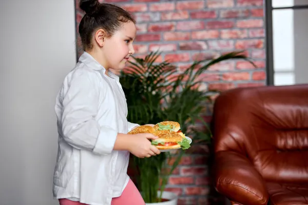 Πλευρική άποψη για βιασύνη κορίτσι παιδί κρατώντας πιάτο με χάμπουργκερ, πρόκειται να παρακολουθήσουν τηλεόραση — Φωτογραφία Αρχείου