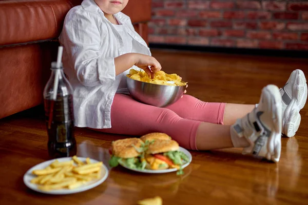 Bijgesneden vet meisje eten chips uit kom terwijl zitten op de vloer in de woonkamer, zijaanzicht — Stockfoto