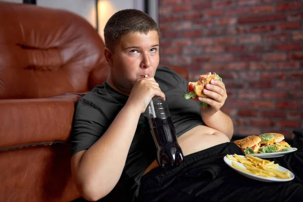 Bambino in sovrappeso con fast food sul pavimento vicino al divano a casa, mangiare cibo spazzatura — Foto Stock