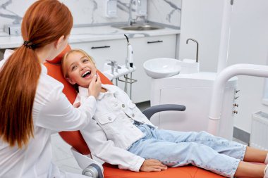 Çocuk dişçi dişlerini kontrol ederken ağzı açık bir şekilde diş kliniğinde kanepede oturuyor.