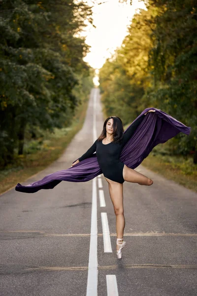 Ballerina v černém obleku a baletní boty tančí na silnici asfalt, skáče a ohyby — Stock fotografie