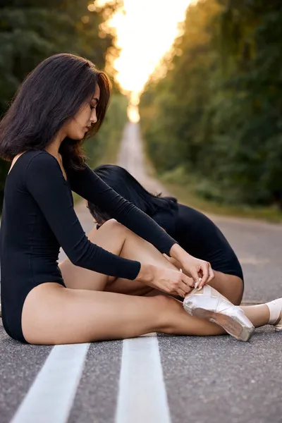 Boční pohled na brunetku samice vázání špičaté boty sedí na silnici, v lese — Stock fotografie