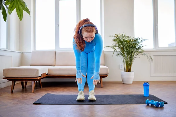 Kilolu kızıl saçlı kadın evde esneme egzersizi yapıyor, spor paspası üzerinde duruyor. — Stok fotoğraf