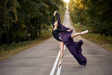 İnce bir balerin yolda ormanda egzersiz yapıyor. Yarı saydam kumaşlı kadın dansçı.