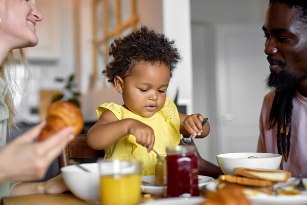 Όμορφη πολυεθνική οικογένεια με το παιδί να παίρνει πρωινό στο σπίτι στη φωτεινή ζεστή κουζίνα. — Φωτογραφία Αρχείου