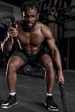 Formda bir zenci modern karanlık spor salonunda savaş halatları kullanarak antrenman yapıyor..