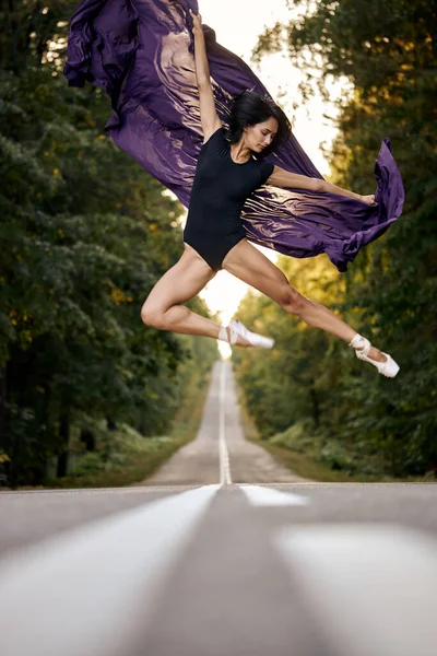 Ballerina v černém obleku a baletní boty tančí na silnici asfalt, skáče a ohyby — Stock fotografie