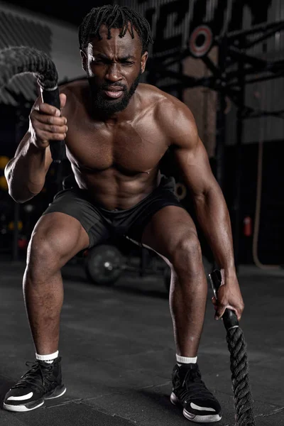 Ajuste hombre negro haciendo entrenamiento de fuerza usando cuerdas de batalla en moderno gimnasio oscuro. —  Fotos de Stock