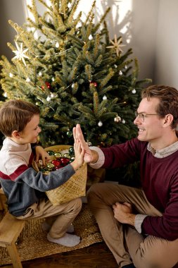 Dost canlısı genç adam küçük oğluna Noel ağacını oyuncaklarla süslemesinde yardım ediyor.