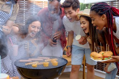 Bir grup arkadaş barbekü ızgarasında yemek yapıyor, çeşitli insanlar piknikte