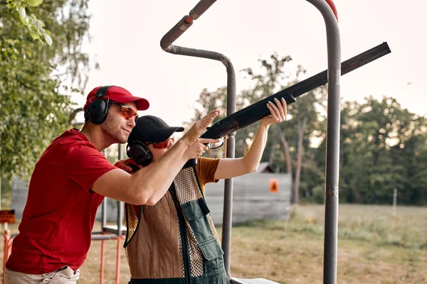 Bonito caucasiano macho no fone de ouvido está ensinando mulher a usar rifle, arma ao alcance — Fotografia de Stock